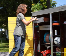 . The image shows a person interacting with a garden storage unit that contains various items. The storage unit is outdoors and has shelves with objects like a hose reel, a soccer ball, a toy car, and other gardening or sporting equipment. There are plants on top of the unit, and the person appears to be arranging or retrieving something from a shelf. The background includes greenery and trees.