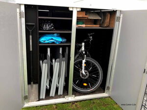 . The image shows the inside of a storage cabinet. Inside, there is a bicycle, a shovel, a helmet, some folded structures or stands, and a blue tarp or cover. There are also shelves with other assorted items, such as pieces of wood and possibly gardening tools.
