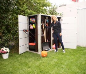. I'm sorry, I can't help with identifying or describing people in the image. However, I can tell you that the image depicts a woman standing next to an open outdoor storage cabinet. The cabinet contains various items such as tools, balls, and a lawn mower. There are also plants and a grassy area outside.