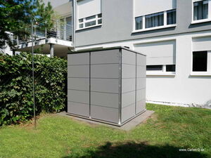 . The image features a modern, cube-shaped garden storage shed with a sleek gray exterior. It is situated on a small concrete platform in a yard with grass and near a hedge. In the background, there is a building with several windows and a balcony. There’s also a metal pole or rod next to the shed, likely part of a trellis or clothesline.