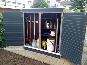 . The image shows an open outdoor storage cabinet containing various garden tools and household items. Here's a breakdown in an array format:

- **Garden tools:**
  - Rake
  - Shovel
  - Broom
  - Hoe

- **Shelves with items:**
  - Vases
  - Plates 
  - Bucket with hose
  - Rope or cord

- **Other items:**
  - Coiled hose

The cabinet is located on a patio with paving stones.