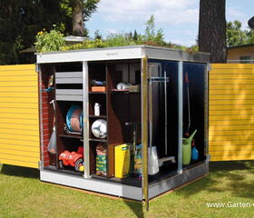 . This image shows a garden storage unit with open doors, revealing various gardening and outdoor tools. Inside, there are items like a hose reel, a soccer ball, garden tools, and other supplies. The roof of the storage unit has plants growing on it, indicating a green roof design.
