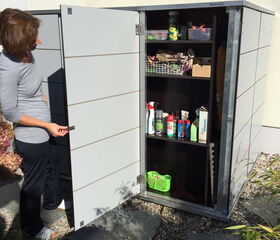 . The image shows a woman standing beside an outdoor storage cabinet with open doors. Inside the cabinet, there are multiple shelves holding various items such as cleaning supplies, gardening tools, and containers. The area around has stone paving and some plants.
