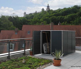 . This image shows a rooftop scene with a small green structure, possibly a storage unit or shed, that is open and contains various tools. The rooftop features a garden area with plants and decorative pots. In the background, there are red-tiled roofs and a wooded area with a large building on a hill. The scene is tranquil, and it appears to be in a well-maintained urban setting.
