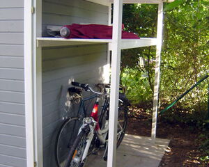 The image shows an outdoor storage area with a partially covered roof. Inside, there are two bicycles parked. Above the bicycles, there's a shelf holding rolled-up items, possibly fabrics or mats. The structure is surrounded by greenery.