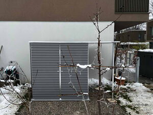 . Ein graues Gartenschrank vor einer Wand, mit verschneiten Bäumen und einem Wasserschlauch auf der linken Seite.
