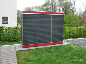 . The image shows a large storage unit or shed with a modern design. It has a black horizontal panel exterior with red accents on the top and bottom. The shed is located outdoors on a lawn, surrounded by hedges and trees, and there's a paved pathway leading up to it.