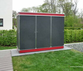 . The image shows a garden storage unit with a sleek, modern design. It features dark panels with metallic edges, and a red base. The unit is situated on a paved area in a garden, with grass, shrubs, and trees in the background. There is also a building with a window to the left.