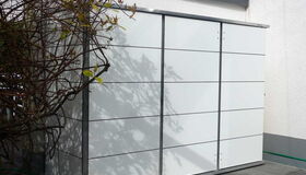 . The image shows an outdoor storage shed with a sleek, modern design. It has a flat roof and smooth, panel-like surfaces. The shed is positioned against a wall, and there are tree branches with leaves casting shadows on its surface. The setting appears to be a patio or a paved area.