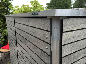 . Holzverkleidetes Gartenmodul mit Metallecken, darauf steht „Garten[Q]“. Im Hintergrund Pflanzen und Bäume.