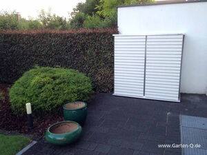 . The image shows a garden scene with a white horizontal slatted storage unit against a wall. In front of it, there are some round, empty green pots on a paved surface. To the left, there is dense greenery and a hedge. There’s also a small outdoor light fixture visible near the plants.