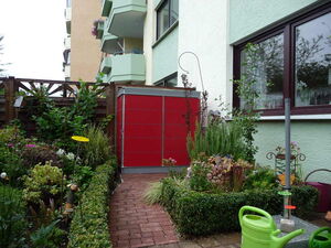 . The image shows a garden space next to a residential building. There's a red storage unit, a variety of plants and flowers, a tall metal stand, a wooden fence, and a green watering can. The building has a light green facade with windows and balconies.