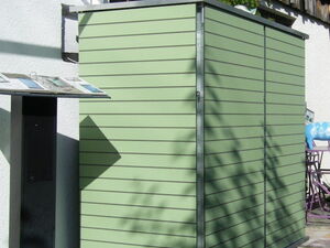 . The image shows a green outdoor storage shed with horizontal panels. It’s positioned against a white wall, and there are some plants and wooden elements nearby. There is also a black stand or information board next to the shed.