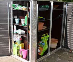 . The image shows an outdoor storage cabinet with open doors. Inside, there are shelves holding various gardening supplies, such as:

- Bags of soil or fertilizer
- Plant pots
- Gardening tools
- A spray bottle
- Boxes likely containing gardening supplies
- A pink watering can

The storage unit appears to be organized for easy access to gardening materials.