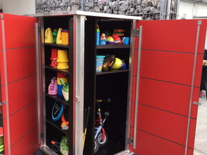 . The image shows an open cabinet with shelves containing various items. Here's an array of what's visible:

1. **Top Shelf:**
   - Small watering cans
   - Colorful toy buckets

2. **Middle Shelf:**
   - Various plastic toys
   - A stuffed toy

3. **Bottom Shelf:**
   - More toys and equipment
   - Small bike or tricycle

4. **Inside the Cabinet:**
   - Various sports equipment (e.g., balls, possibly a hoop or rope)
   - Items organized for easy access

The cabinet itself is red with metal framing and seems to be located outdoors.