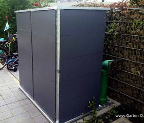 . The image shows a sleek, modern garden storage unit with a rectangular shape, set on a patio. This storage unit has a minimalist design with smooth, dark panels. Next to it on the left are parked bicycles. To the right, there's a wire fence filled with rocks, which resembles a gabion wall. There's also a green pipe with a protective cage near the unit. The ground is paved with square tiles, and there's vegetation visible around the area.