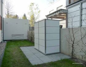 The image shows a small, well-maintained backyard space that includes a large white storage unit or shed on a patio made of square tiles. The area is bordered by a fence and surrounded by grass, with small trees or bushes lining the sides. There is also a flat bench positioned against one of the walls.