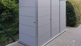 . Ein grauer Gartenschrank aus Metall steht auf einer gepflasterten Fläche, umgeben von Bäumen und einem Zaun.