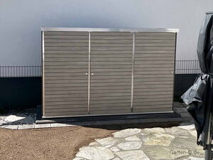 . Ein Metallschuppen steht im Freien auf einem gepflasterten Bereich, daneben ein zusammengeklappter Sonnenschirm.