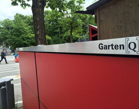 . Ein großer, roter "Garten[Q]" Schrank im Freien, Baum und Leute im Hintergrund.