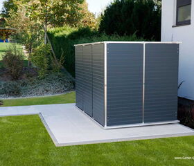 . The image shows a modern, compact metal storage unit or garden shed placed on a tiled patio in a landscaped garden. It's situated next to a building with an exterior wall. The surrounding garden area has neatly trimmed grass, shrubs, and a few trees, creating a tidy and organized outdoor space.