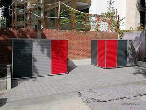 . The image shows two storage units with a modern design positioned on a paved area. Both units have a combination of dark gray, light gray, and red panels. They are placed in front of a brick wall, and there's a large structure with netting and plants behind the wall. A piece of fabric or covering material is laid out on the ground in front of the units.