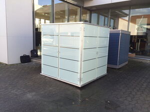 . Ein gläserner Kasten steht draußen vor einem Gebäude mit großen Fenstern, daneben zwei Aktenkoffer.