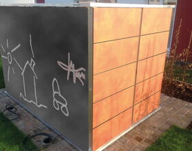 . The image shows a rectangular structure with two distinct sides. One side is a chalkboard surface featuring simple chalk drawings, including a sun, a house, and some abstract shapes. The other side is made of wooden panels with a horizontal slat design. The structure is placed on a paved surface with some grass and landscaping around it.