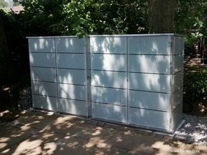 . The image shows a metallic structure with a sleek design, likely a storage unit or cabinet. This structure is situated outdoors, surrounded by trees and foliage, and is placed on a stone or paved surface. It appears to have multiple compartments with horizontal lines, indicating separate sections or doors for storage.
