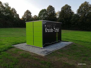 . Ein grünes Gartenhaus mit einer Kreidetafel, auf der "Kreide-Tafel" und Zeichnungen stehen, steht auf einer Wiese.