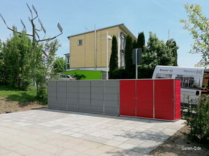 . Ein moderner Gartenschrank mit grauen und roten Paneelen steht auf einem Plattenboden neben einem Haus.