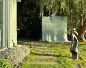 . Ein Garten mit moderner Skulptur, Gehwegplatten und Bäumen, im Hintergrund eine Wand oder Struktur.