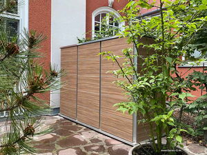 . The image shows a modern garden storage shed with a wood-like finish. It is placed against an exterior wall of a house, surrounded by greenery, including potted plants and a tree. The house has a red and white exterior with windows. The ground is covered with stone pavement.