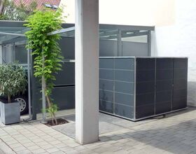 . The image shows an outdoor area with modern design elements including:

1. A metal structure, possibly a carport or storage area.
2. A gray storage unit or enclosure.
3. Paved flooring.
4. Green plants, including a small tree in a planter and climbing vines on the metal structure.
5. A partial view of a parked car under the structure.

The setting appears to be a modern residential or commercial outdoor space.