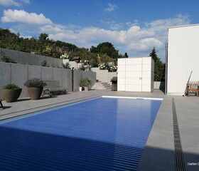 Ein moderner Garten mit Pool, Betonwänden, Pflanzen in Töpfen, Bänken und einem weißen Gebäude.