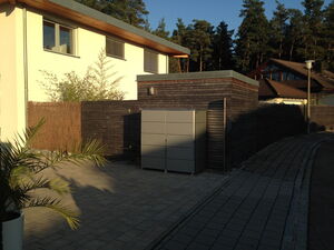 . The image shows a residential area with a modern house. The house has a white exterior with wooden trim. There is a paved driveway or walkway leading up to it. To the right, a wooden fence lines the property, and there is a small, gray, box-shaped structure near the entrance. Trees are visible in the background, and a potted plant is in the foreground. The area is well-lit, indicating it might be during daylight hours.