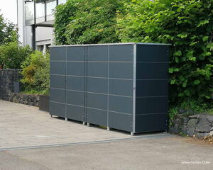The image shows a modern outdoor storage unit or cabinet placed alongside a paved area, likely in a garden or driveway setting. The structure is surrounded by greenery including bushes and trees, and there is a stone wall to the left. The storage unit has a sleek, modular design with a dark color and metal framework.