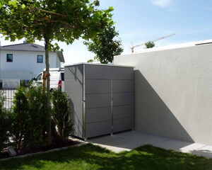 . The image shows a small outdoor area featuring a metal storage cabinet against a light-colored wall. There’s a tree with green leaves on the left, some shrubs, and a patch of grass in the foreground. In the background, there is a white building and a crane visible, along with a parked white vehicle.