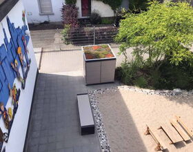 . Ein Garten mit Sitzbank, Wandkunst, Baum, Sandfläche und bepflanztem Hochbeet aus der Vogelperspektive.