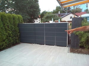 . The image shows an outdoor scene with a tiled patio area. There is tall greenery on the left, likely trees or shrubs, and dark-colored outdoor cabinets or panels along the back. A person’s arm is extending from the right side, pointing towards the cabinets or panels. In the background are trees and some buildings.