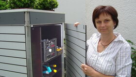 . The image shows a person standing next to an open storage unit or cabinet. The cabinet has shelves with various tools or items, and there are potted plants on top. The person is holding the door open.