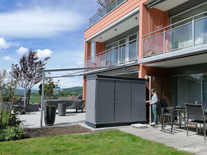 . Eine Terrasse mit Möbeln, Gartenhaus und einer Person vor einem modernen Gebäude mit Glasgeländern und Balkon.