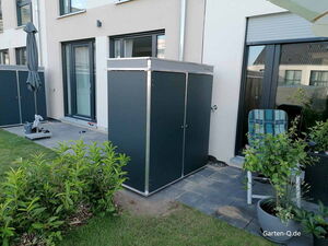 . Ein Garten-Lagerschrank auf einer Terrasse, umgeben von Pflanzen, Stühlen und einem Sonnenschirm.