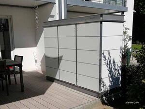 . Ein Gartenschrank auf einer Terrasse neben einem Tisch mit Stühlen, umgeben von Pflanzen und Sonnenschein.