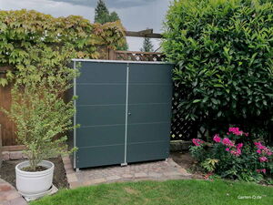 . Ein graues Metallschrank im Garten mit Pflanzen, Hecke und Blumen drumherum.
