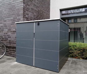 . Ein grauer Metallschrank neben einer Hauswand mit Fahrradreifen im Vordergrund.