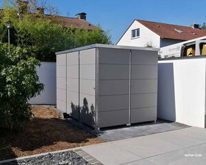 . Garten mit modernem grauen Geräteschrank vor einem weißen Zaun, umgeben von Büschen und einem Haus im Hintergrund.