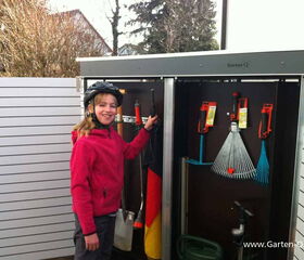 . The image shows a person standing next to an outdoor storage unit containing various garden tools. The tools inside include a rake, a broom, and some other implements neatly organized. The person is wearing a helmet and a pink jacket, smiling at the camera. The storage unit is located outside, with some fencing and trees visible in the background.