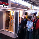 The image shows a group of people observing a display at an indoor event, possibly a trade show or exhibition. The display appears to feature a storage shed with open doors revealing garden tools or equipment inside.