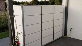 . Ein moderner Gartenschrank auf einer Terrasse mit Pflanzen und Stuhl, überdacht durch Glasdach.